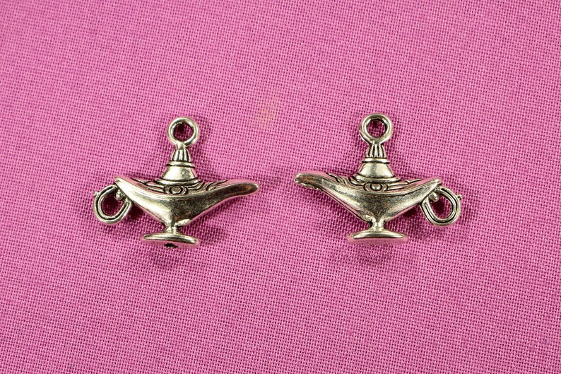 May include: Two silver-toned genie lamp charms with intricate details. The lamps have a rounded base and a spout with a loop handle. The charms are on a pink fabric background.