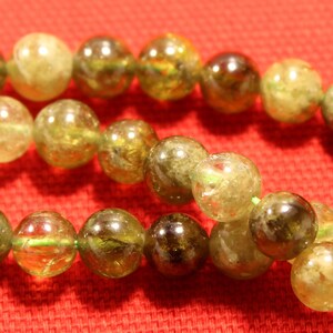 May include: A strand of green gemstone beads with a shiny, translucent finish. The beads are round and have a slightly irregular shape. The beads are strung on a clear thread.