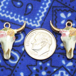 May include: Three gold-tone metal charms shaped like bull skulls with floral designs. The charms are on a blue and white patterned fabric background.