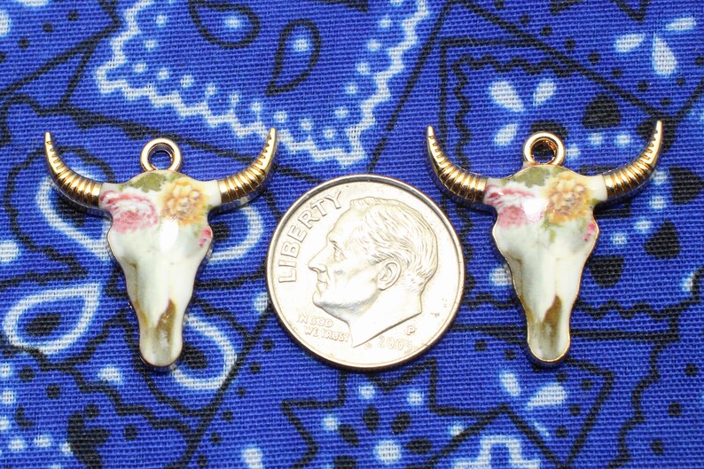 May include: Three gold-tone metal charms shaped like bull skulls with floral designs. The charms are on a blue and white patterned fabric background.