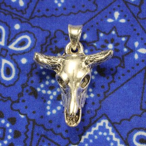 May include: Silver pendant in the shape of a bull skull with horns. The pendant is on a blue and white patterned fabric background.