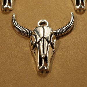 May include: Silver-toned metal bull skull charm with detailed horns and a skull design. The charm has a loop at the top for attaching to a necklace or bracelet.