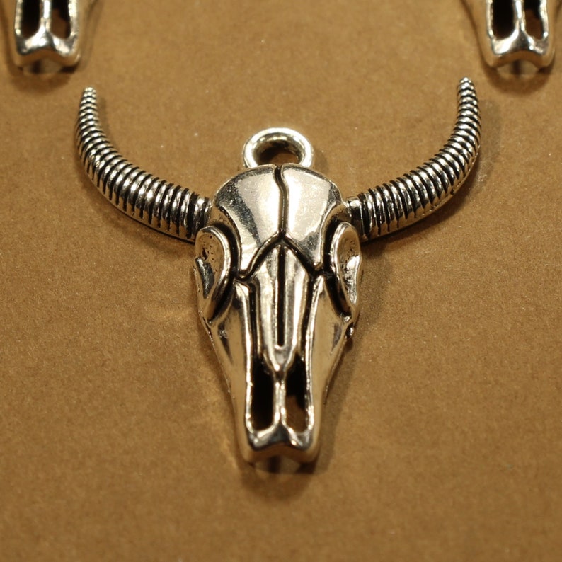 May include: Silver-toned metal bull skull charm with detailed horns and a skull design. The charm has a loop at the top for attaching to a necklace or bracelet.