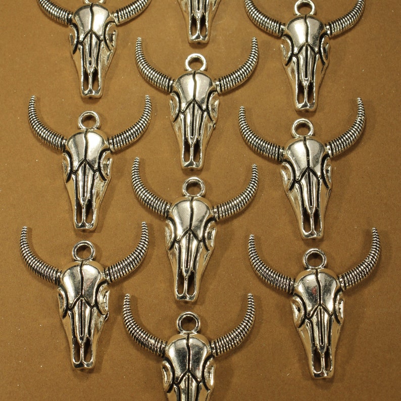 May include: A collection of silver-toned metal charms shaped like bull skulls. Each charm has intricate details and a loop for attaching to a necklace or bracelet.