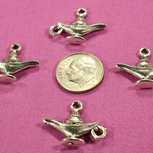 May include: Four silver-toned genie lamp charms, each with a detailed design, are arranged on a pink fabric background. A US dime coin is placed in the center for size comparison.