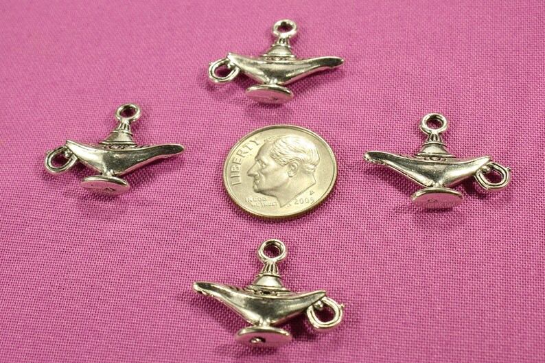 May include: Four silver-toned genie lamp charms, each with a detailed design, are arranged on a pink fabric background. A US dime coin is placed in the center for size comparison.
