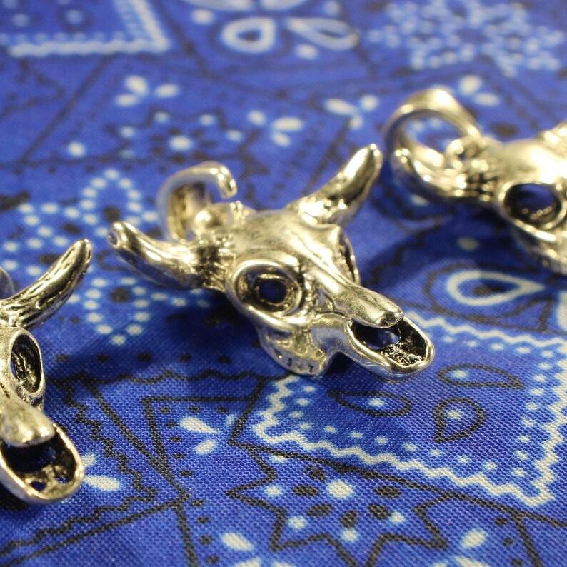 May include: Three silver skull charms with horns, resembling a bull's skull, are displayed on a blue and white patterned fabric.