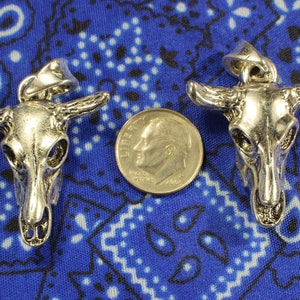 May include: Two silver-toned bull skull pendants on a blue and white patterned fabric background. The pendants are approximately the size of a US dime.