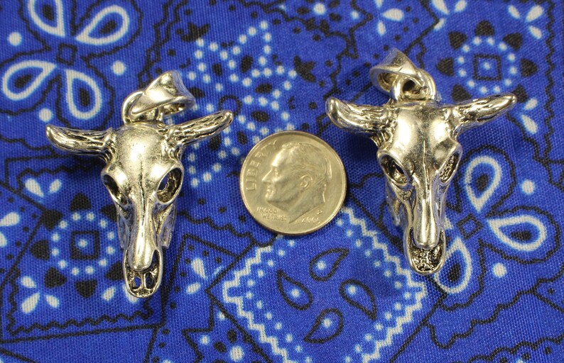 May include: Two silver-toned bull skull pendants on a blue and white patterned fabric background. The pendants are approximately the size of a US dime.