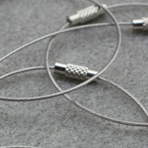 May include: Close-up of three silver keyrings with metal loops and crimped ends. The keyrings are made of braided steel cable.