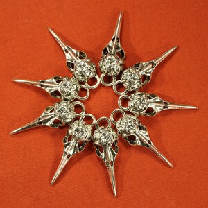 May include: Eight silver bird skull charms with intricate details, arranged in a star shape on a red background.