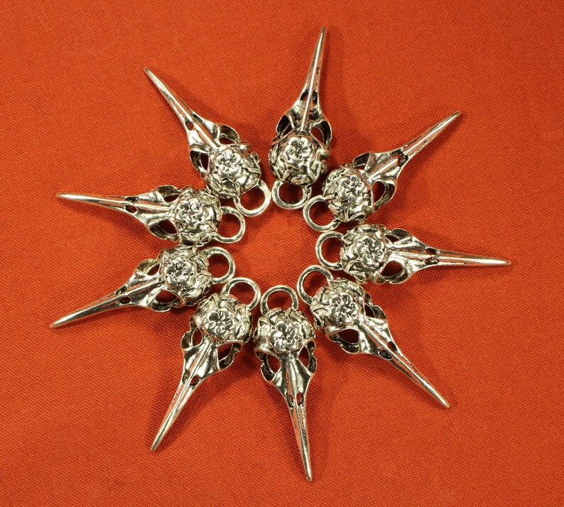 May include: Eight silver bird skull charms with intricate details, arranged in a star shape on a red background.