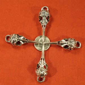 May include: Four silver-toned metal bird skull charms with floral details. The charms are arranged in a cross shape with a silver-toned coin in the center.