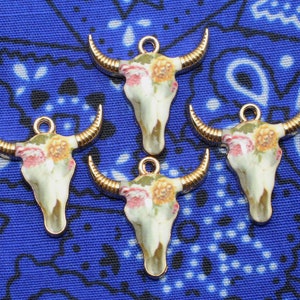 May include: Four gold-tone bull skull charms with floral designs. The skulls are white with pink and yellow flowers.