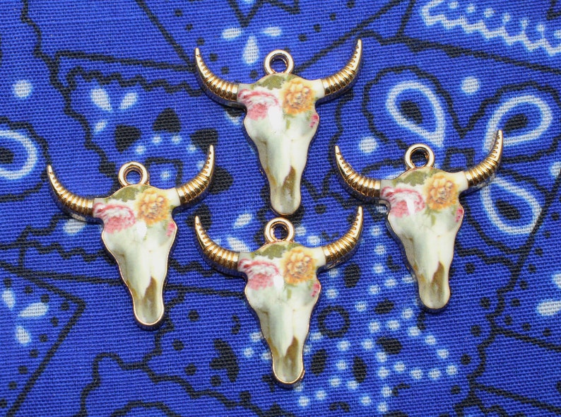 May include: Four gold-tone bull skull charms with floral designs. The skulls are white with pink and yellow flowers.
