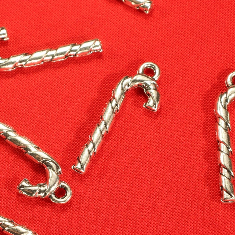 Op de afbeelding: Zilverkleurige snoepstokcharms met een gedraaid ontwerp. De charms bevinden zich op een rode achtergrond.