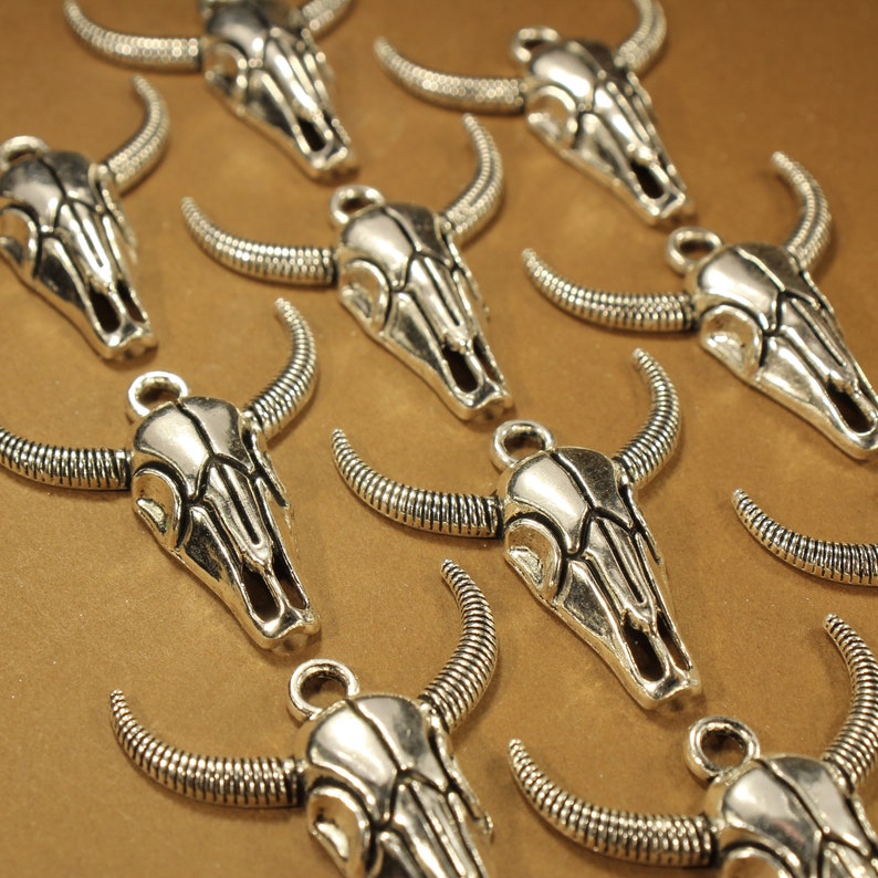 May include: A collection of silver-toned metal charms shaped like bull skulls. The charms have intricate details and are perfect for jewelry making or crafting.