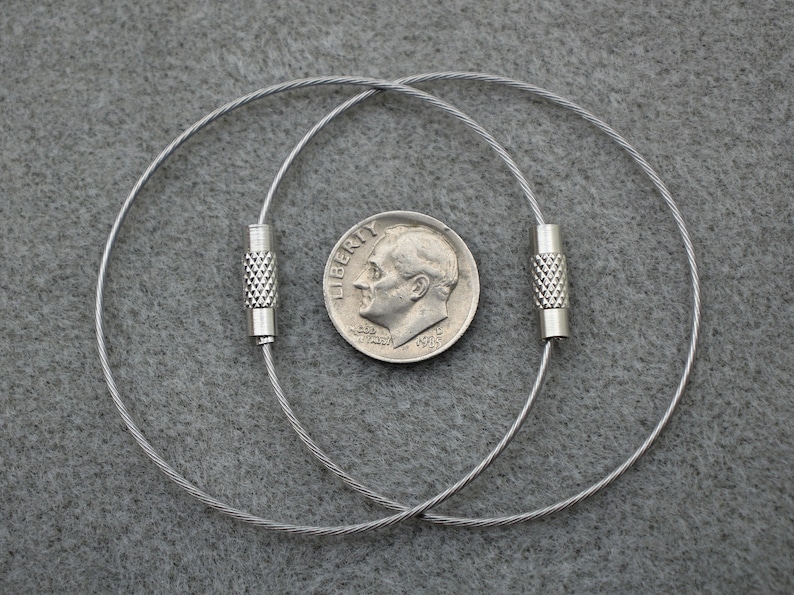 Puede incluir: Dos llaveros plateados con un dise&ntilde;o de cable y tapas de extremo plateadas. Los llaveros est&aacute;n dispuestos sobre una superficie gris texturizada.