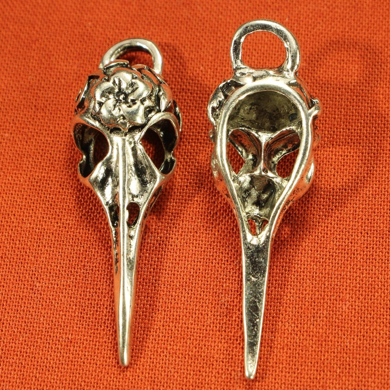 May include: Two silver bird skull pendants with intricate details. One pendant has a floral design on the top, while the other is open and shows the inside of the skull.