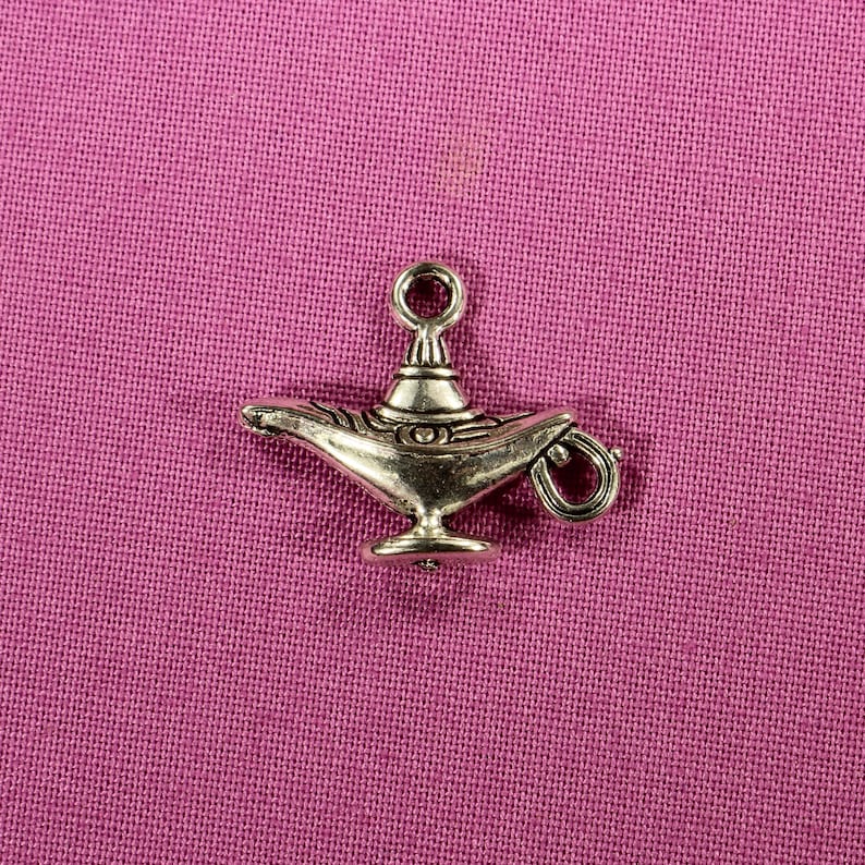 May include: A silver charm in the shape of an Aladdin's lamp. The lamp has a detailed design and a small loop at the top for attaching to a necklace or bracelet.