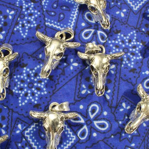 May include: A collection of silver-toned bull skull pendants on a blue bandana background. The pendants have intricate details and are perfect for jewelry making or crafting.