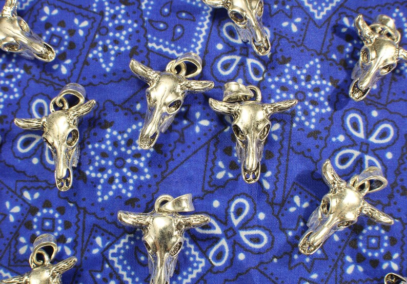 May include: A collection of silver-toned bull skull pendants on a blue bandana background. The pendants have intricate details and are perfect for jewelry making or crafting.