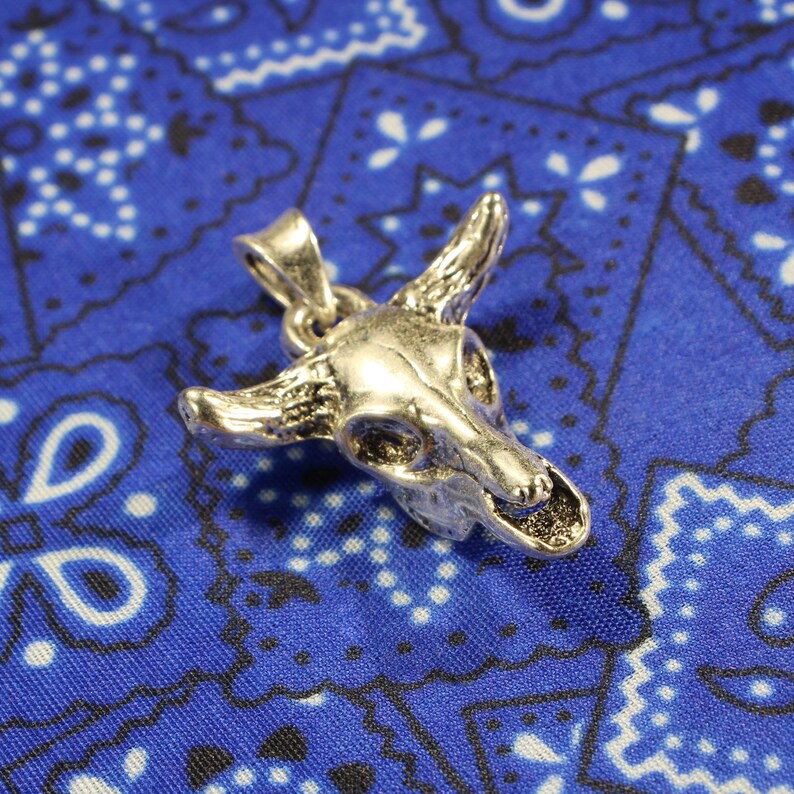 May include: Silver-toned pendant in the shape of a bull skull. The pendant is on a blue and white patterned fabric background.