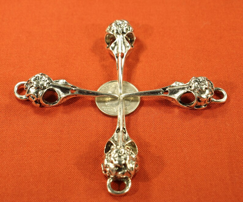 May include: Four silver bird skull charms arranged in a cross shape. The charms are detailed with intricate carvings and have a loop at the top for attaching to jewelry.