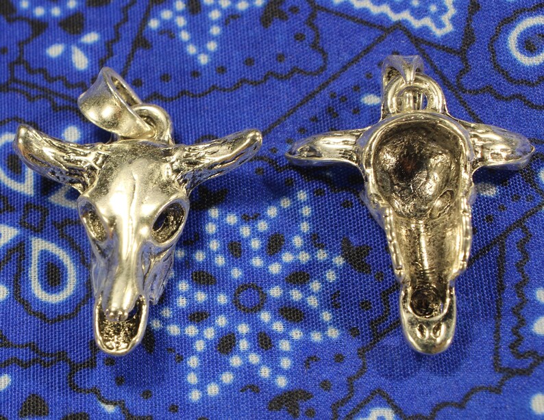 May include: Two silver skull pendants with a bull's head design. The pendants are on a blue and white patterned background.