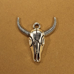 May include: Silver-toned metal charm in the shape of a bull skull with a loop at the top for attaching to a necklace or bracelet.