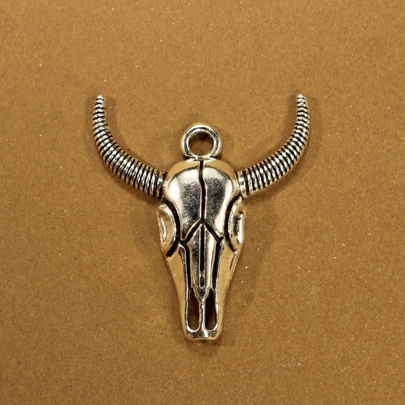 May include: Silver-toned metal charm in the shape of a bull skull with a loop at the top for attaching to a necklace or bracelet.