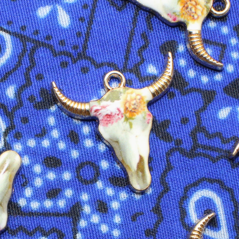 May include: A white porcelain bull skull charm with gold accents and a floral pattern. The charm features a detailed design of roses and other flowers in shades of pink, yellow, and green.
