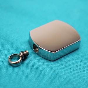 May include: A shiny, silver-colored pendant with a rounded rectangular shape. The pendant has a small hole and a separate screw-in loop. The background is a solid teal color.