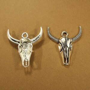May include: Two silver-toned metal bull skull charms. One charm has horns that curve upward and the other has horns that curve outward.