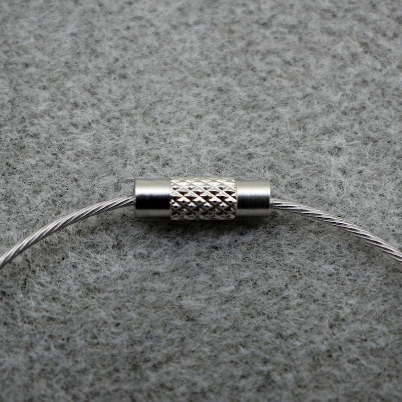 Puede incluir: Un llavero de metal plateado con un bucle de cable y un conector de metal texturizado.