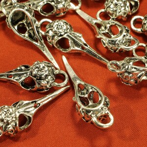 May include: A collection of silver-toned metal charms shaped like bird skulls with intricate floral details. The charms have a loop at the top for attaching to jewelry or other crafts.