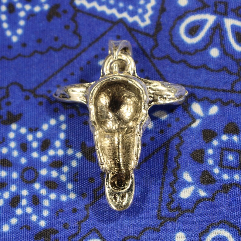 May include: Silver skull pendant with intricate details. The skull is facing forward with the back of the pendant visible. The pendant is on a blue and white patterned fabric.