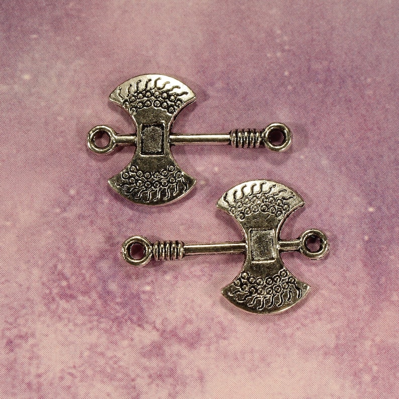 May include: Two silver axe-shaped charms with intricate designs. The charms have a loop at the top for attaching to a necklace or bracelet.