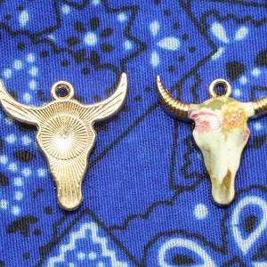 May include: Two gold-toned bull skull charms with different designs. One charm is plain and the other has a floral pattern.
