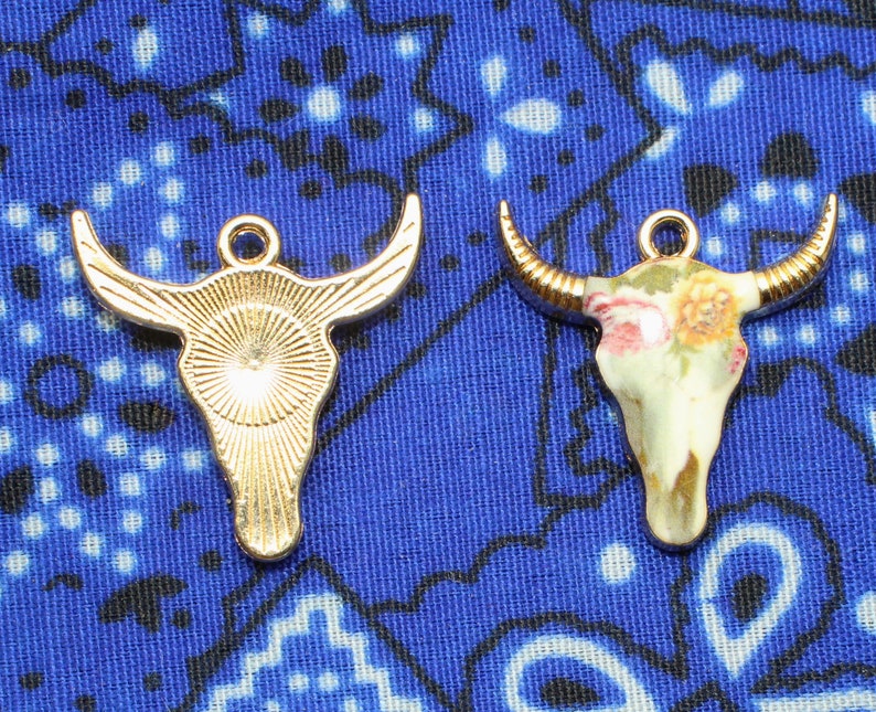 May include: Two gold-toned bull skull charms with different designs. One charm is plain and the other has a floral pattern.