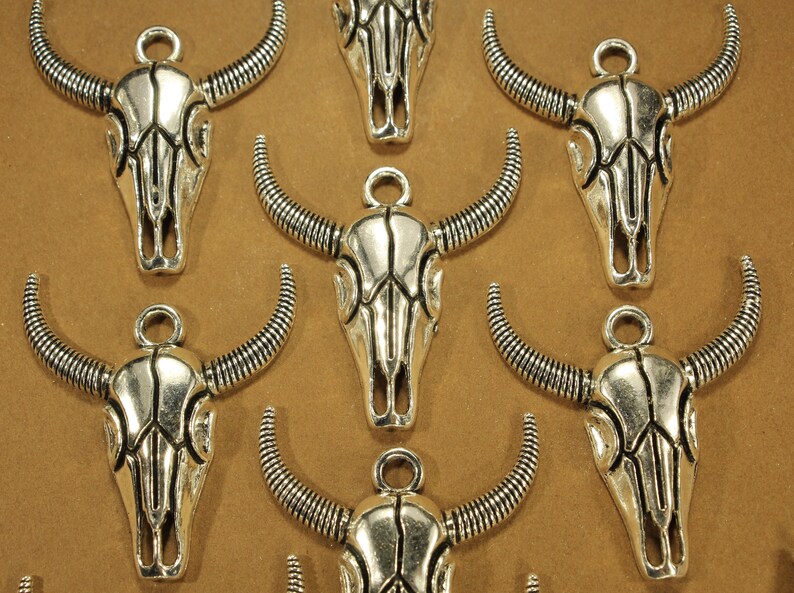 May include: Nine silver-toned bull skull charms with intricate details. Each charm features a detailed skull with horns and a loop for attaching to jewelry or crafts.