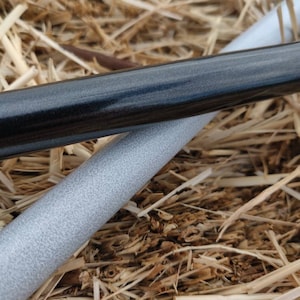 May include: Two long, cylindrical rods, one black and one silver, are lying on a bed of straw. The black rod has a smooth, glossy finish, while the silver rod has a textured surface.