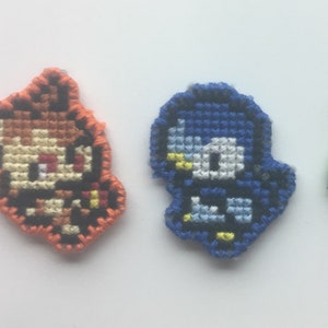 May include: Three embroidered patches featuring cartoon characters. The first patch is orange and features a character with brown hair and a red shirt. The second patch is blue and features a character with blue hair and a blue shirt. The third patch is green and features a character with brown hair and a yellow shirt.