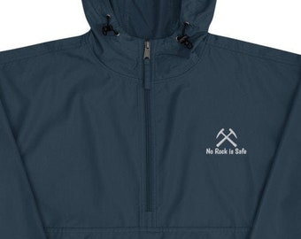 Puede incluir: Una chaqueta cortavientos azul oscuro con capucha, cierre de cremallera y un logotipo bordado en blanco que dice "No Rock Is Safe" con dos picos cruzados. La chaqueta tiene un logotipo de Champion en la manga izquierda.
