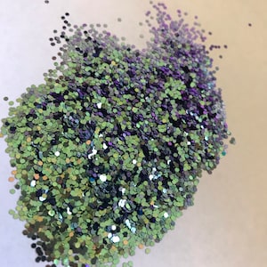 Puede incluir: Un montón de purpurina iridiscente verde y morada. La purpurina tiene forma de pequeños hexágonos.