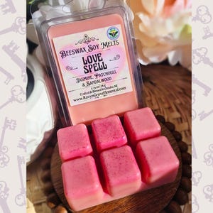 May include: Six pink beeswax soy wax melts in a clear plastic container. The label reads "Beeswax Soy Melts Love Spell Jasmine, Patchouli, & Sandalwood 2.75 oz / 78 g Crafted & Blessed in Omaha, NE www.RavynGroveElemental.com"
