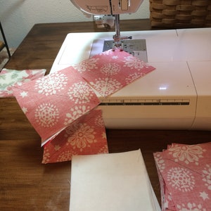 May include: A sewing machine with a white body is shown with pink and white patterned fabric squares. The fabric squares are in various stages of being sewn together. A wooden table and a basket with floral fabric are in the background.