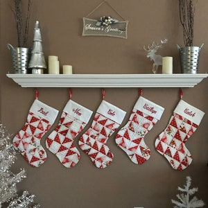 May include: A festive Christmas display featuring five stockings with red and white geometric patterns and embroidered names. The stockings hang below a shelf with decorations, including candles and a sign that reads "Season's Greetings".