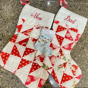 May include: Two Christmas stockings with red and white geometric patterns, embroidered with "Mom" and "Dad" in red. A smaller stocking with a gnome and snowman pattern is nestled between them. Each stocking has a white cuff and red ribbon loop.