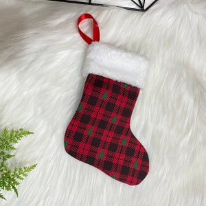 May include: Miniature Christmas stocking with a red and black plaid pattern and small green Christmas trees. The top of the stocking is trimmed with white faux fur and has a red ribbon loop for hanging.